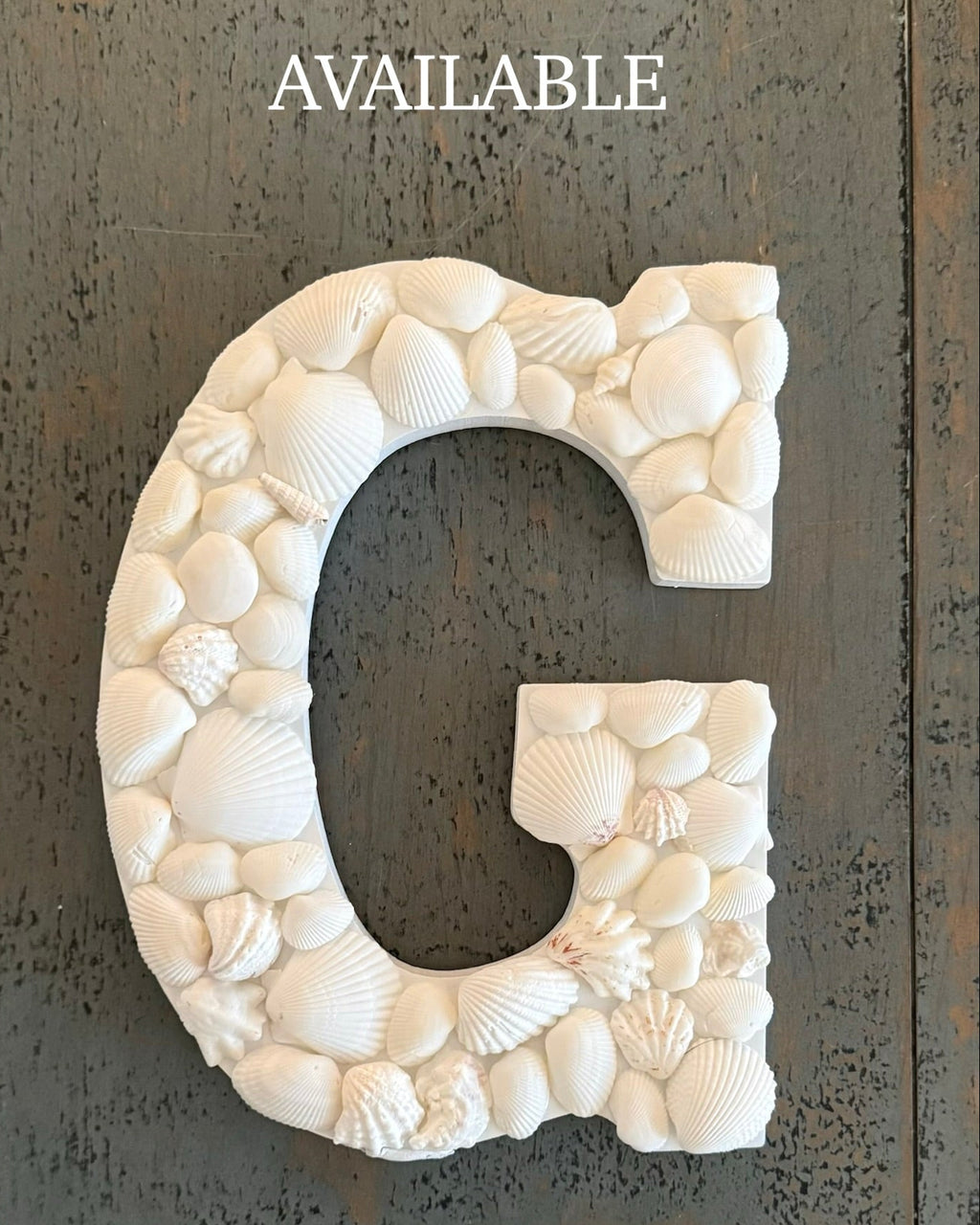 Decorative letter 'G' made of seashells on a textured surface with 'AVAILABLE' text above.