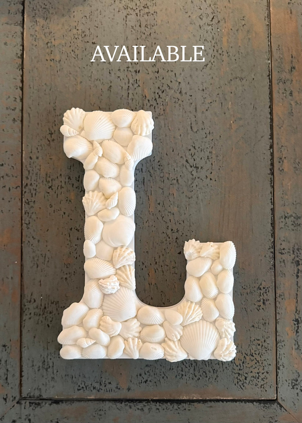 Decorative letter 'L' made of seashells on a textured surface with 'AVAILABLE' text above.