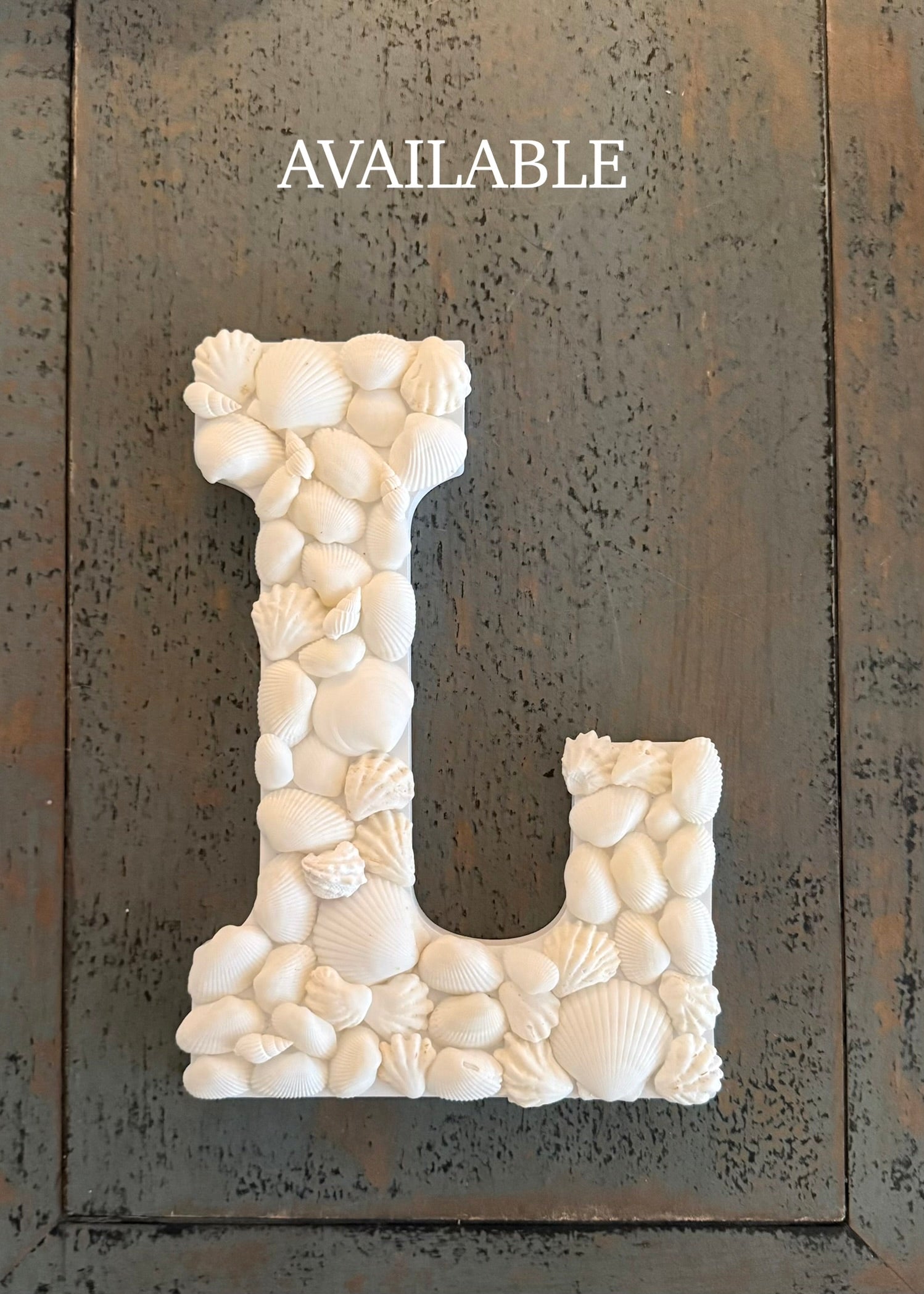 Decorative letter 'L' made of seashells on a textured surface with 'AVAILABLE' text above.