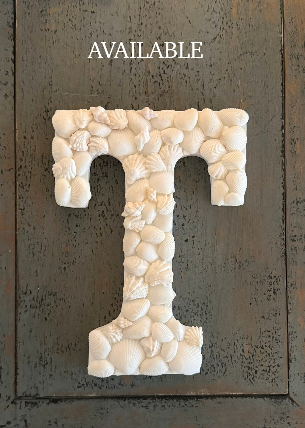 Decorative letter 'T' made of seashells on a textured dark background with 'AVAILABLE' text above.