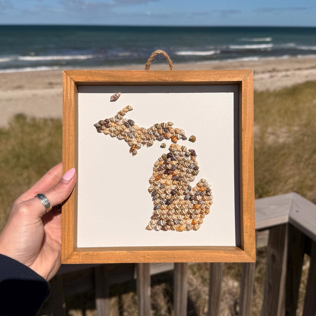 Framed artwork of a map of Michigan held by a person on a beach with ocean and sky in the background.