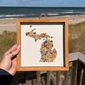 Framed artwork of a map of Michigan held by a person on a beach with ocean and sky in the background.